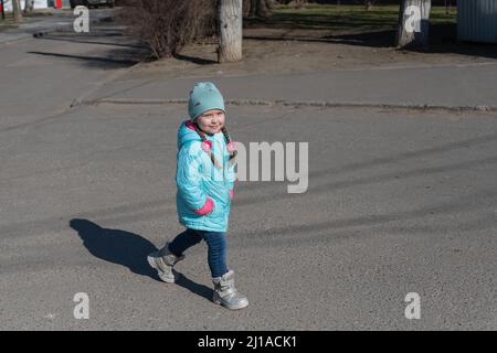 Ein Kind geht auf einem asphaltierten Bürgersteig. Ein fünfjähriges Mädchen mit den Händen in den Taschen. Ein sonniger Frühlingstag. Seitenansicht. Stockfoto