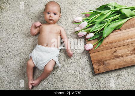 Niedliches kleines Baby liegt auf dem Boden neben dem Blumenstrauß der Tulpen Stockfoto