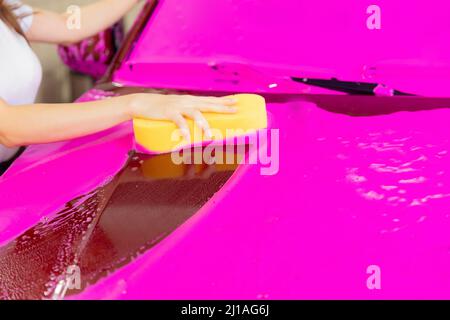Halten Sie den gelben Schwamm mit der Hand über das Auto, um den rosa Schaumstoff-Hintergrund zu waschen. Stockfoto