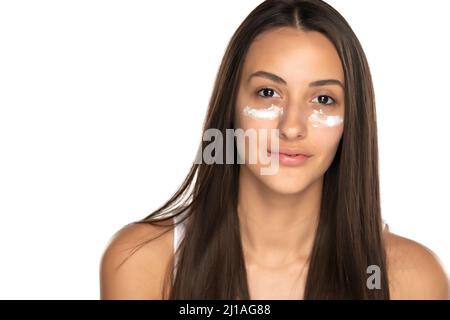Eine junge, schöne Frau, die mit Gesichtscreme unter ihren Augen auf weißem Hintergrund posiert