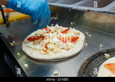 Koch bereitet Pizza auf einem Metalltisch zu, Nahaufnahme. Nahaufnahme Hand des Chefbäckers in Gummihandschuhen Zugabe von Zutaten zur Pizza in der Küche. Stockfoto