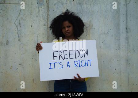 Eine afroamerikanische Frau mit einem Banner in ihren Händen, das sagt, dass Freiheit ein Recht sei. Im Hintergrund graue Wand Stockfoto