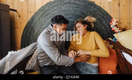 Blick von oben auf ein verheiratetes Paar, das zusammen lacht, während es im Spielbereich ihrer Kinder liegt. Zwei glückliche junge Eltern, die Spaß miteinander haben. Cou Stockfoto