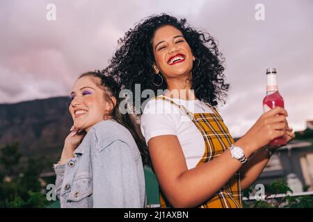 Beste Freunde feiern auf dem Dach. Zwei glückliche Freundinnen, die draußen tanzen und kaltes Bier trinken. Zwei fröhliche junge Frauen, die eine gute Zeit zusammen haben Stockfoto