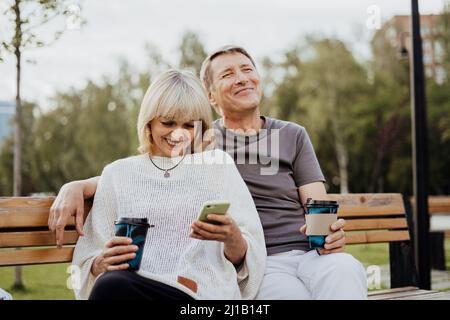 Lachende Erwachsene reifes Paar in der Liebe sitzt auf der Bank mit Telefon im Freien im Park. Ein blonder Kaukasusmann und eine blonde Frau verbringen Zeit miteinander und trinken Stockfoto