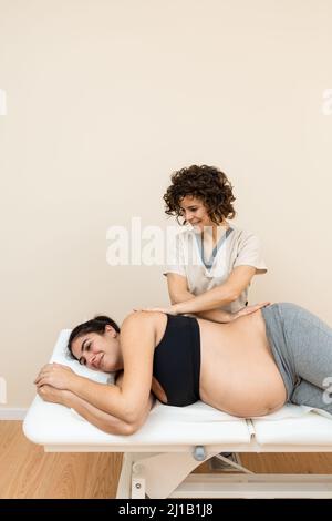 Physiotherapeutin, die eine auf der Seite einer Schwangeren auf einem Massageliegen in einem Gesundheitszentrum liegende Frau massiert Stockfoto