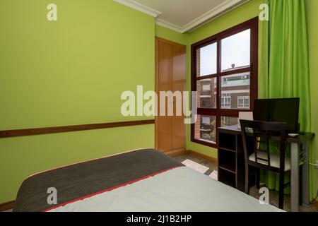 Schlafzimmer mit kleinem dunklen Holzbett mit brauner Decke mit roten Zierleisten, dunklem Schreibtisch mit pc, Fenster mit Aussicht und gestrichenen Wänden in Pistazien-Grün Stockfoto