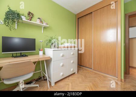 Schreibtisch mit pc auf Holztisch, Kommode mit sechs Schubladen und Einbauschrank mit Schiebetüren aus Eichenholz in einem Raum mit pistaziengrünen Wänden Stockfoto