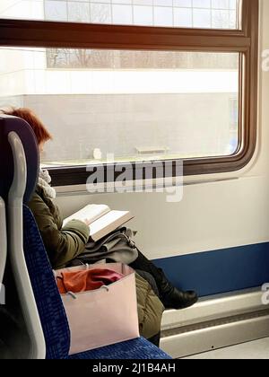 Frau liest auf dem Weg zur Arbeit ein Buch. Leerer Zug fährt früh am Morgen. Ein pinker Beutel mit Habseligkeiten befindet sich auf dem Nachbarsitz. Stockfoto