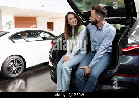 Ein junges Ehepaar in einem Autohaus wählt ein Auto sitzt mit einem Lächeln im Kofferraum eines Crossover Stockfoto