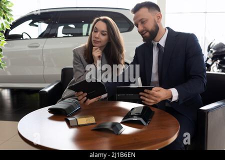 Ein Mitarbeiter eines Autohauses bietet verschiedene Veredelungsmaterialien für ein neues Auto an, das eine junge Frau kauft Stockfoto