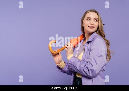 Glückliche junge Frau im Trenchcoat stehend mit orangefarbenem Schirm isoliert auf lila Stockfoto