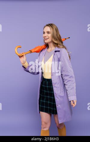 Positive junge Frau im Trenchcoat stehend mit orangefarbenem Schirm isoliert auf lila Stockfoto