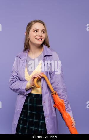 Fröhliche junge Frau im Trenchcoat, die mit einem orangefarbenen Regenschirm auf lila isoliert steht Stockfoto