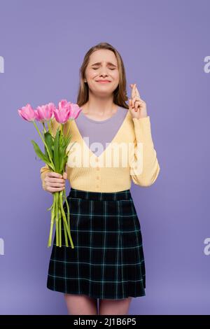 Junge Frau mit rosa Tulpen und stehend mit gekreuzten Fingern isoliert auf lila Stockfoto