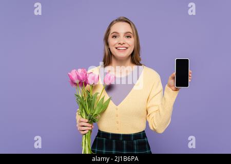 Fröhliche junge Frau mit rosa Tulpen und zeigt Smartphone mit leerem Bildschirm isoliert auf lila Stockfoto