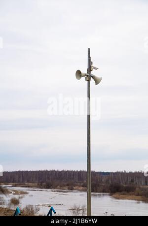 Zwei Lautsprecher-Megaphone auf einem hohen Pol vor dem Hintergrund des Flusses. Frühling, der Schnee schmilzt, es gibt Pfützen von Schlamm und Schlamm ringsum. Tag, bewölktes Wetter, weiches warmes Licht. Stockfoto