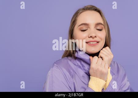 Fröhliche junge Frau mit geschlossenen Augen im Trenchcoat isoliert auf lila Stockfoto