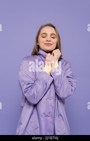 Zufrieden junge Frau mit geschlossenen Augen in Trenchcoat isoliert auf lila Stockfoto