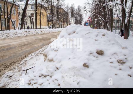 Schneebedeckte große Schneeverwehung in der Nähe der Straße vor dem Hintergrund einer Stadtstraße. Auf der Straße liegt schmutziger Schnee in hohen Haufen. Urbane Winterlandschaft. Wolkiger Wintertag, weiches Licht. Stockfoto