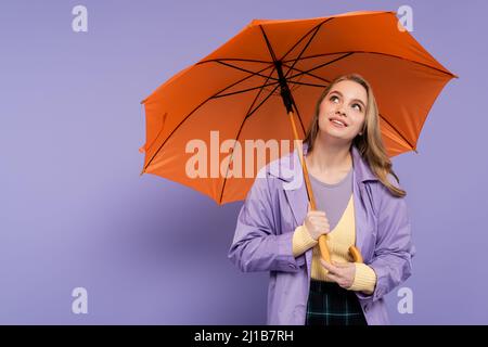 Verträumte junge Frau im Trenchcoat, die unter einem orangefarbenen Schirm auf lila steht Stockfoto