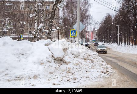 Eine riesige, große Schneeverwehung an der Straße vor dem Hintergrund einer Stadtstraße. Auf der Straße liegt schmutziger Schnee in hohen Haufen. Urbane Winterlandschaft. Wolkiger Wintertag, weiches Licht. Stockfoto