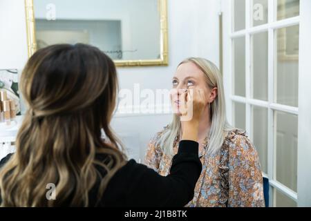 Kosmetologe, der eine blonde Frau in einem Schminkstuhl mit Augen-Make-up bewirbt Stockfoto