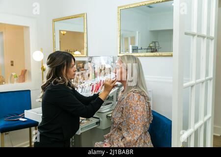 Kosmetikerin, die Foundation Make-up auf eine blonde Frau in einem Schminkstuhl anwendet Stockfoto