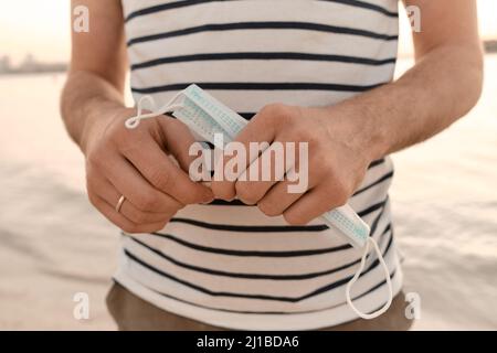 Der Mensch hält nervös eine zerbröckelte medizinische Maske auf dem Meer. Verlassen ist fraglich. Selektiver Fokus. Stockfoto