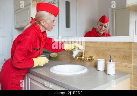 Eine Frau in roter Uniform wischt den Wasserhahn im Badezimmer mit einem Lappen ab. Desinfektion in gelben Gummihandschuhen . Reinigungskonzept. Stockfoto