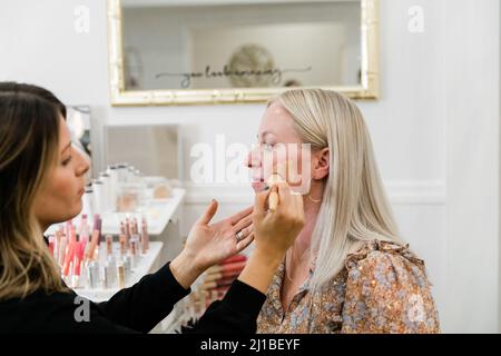 Kosmetikerin, die Foundation Make-up auf eine blonde Frau in einem Schminkstuhl anwendet Stockfoto