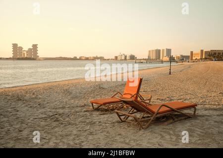 Zwei orangefarbene Sonnenliegen stehen am Meer bei Sonnenuntergang, Dubai VAE. Das Royal Atlantis Dubai auf der Rückseite Stockfoto