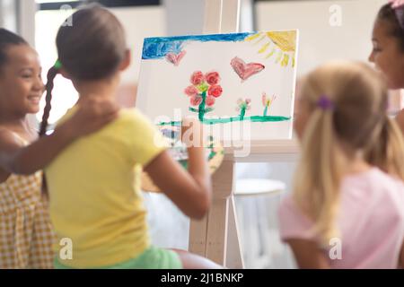 Glückliche multirassische Grundschüler stehen von kaukasischen Mädchen Malerei auf Staffelei in der Klasse Stockfoto