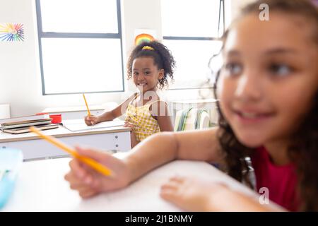 Portrait eines glücklichen Biracial-Mädchens, das auf einem Buch schreibt, während es im Klassenzimmer am Schreibtisch sitzt Stockfoto