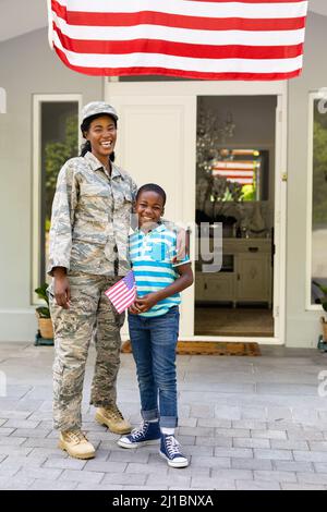 Porträt eines glücklichen afroamerikanischen Soldaten mittleren Erwachsenen mit Sohn, der die amerikanische Flagge vor dem Haus hält Stockfoto