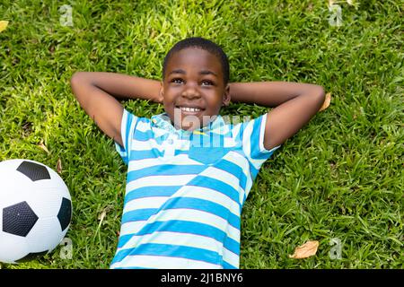 Lächelnder afroamerikanischer Junge, der mit den Händen hinter dem Kopf am Fußballball auf dem Rasen im Hinterhof liegt Stockfoto
