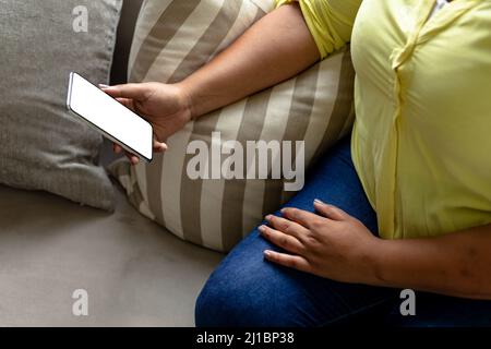Mittelteil einer afroamerikanischen Frau mit mittlerem Erwachsenen, die Smartphone benutzt, während sie zu Hause auf dem Sofa sitzt Stockfoto