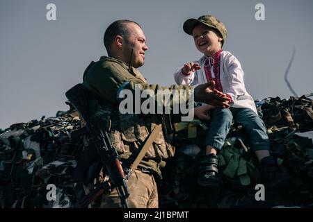 Der Junge im bestickten Hemd sitzt auf Sandsäcken und lacht am Checkpoint in der Nähe des ukrainischen Territorialverteidigungskriegers. Konzept der russischen Militärinvas Stockfoto