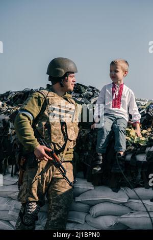 Der Junge im bestickten Hemd sitzt auf Sandsäcken am Checkpoint in der Nähe des ukrainischen Territorialverteidigungskriegers. Konzept der russischen Militärinvasion in Ukra Stockfoto