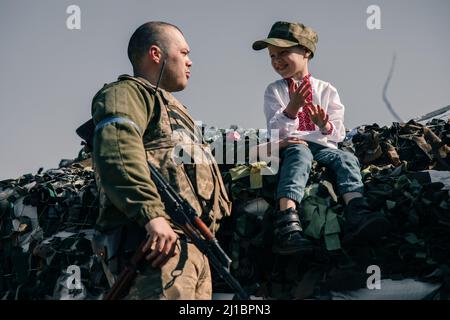 Der Junge im bestickten Hemd sitzt auf Sandsäcken am Checkpoint in der Nähe des ukrainischen Territorialverteidigungskriegers. Konzept der russischen Militärinvasion in Ukra Stockfoto