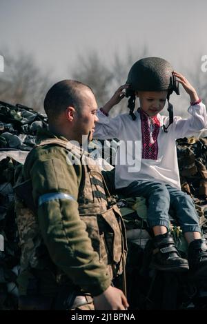 Der Junge im bestickten Hemd sitzt auf Sandsäcken am Checkpoint in der Nähe des ukrainischen Territorialverteidigungskriegers. Konzept der russischen Militärinvasion in Ukra Stockfoto