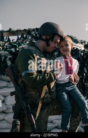 Der Junge im gestickten Hemd sitzt in der Nähe des ukrainischen Territorialverteidigungskriegers am Kontrollpunkt vor Sandsäcken-Hintergrund. Konzept des russischen Militärs Stockfoto