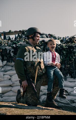 Der Junge im gestickten Hemd sitzt in der Nähe des ukrainischen Territorialverteidigungskriegers am Kontrollpunkt vor Sandsäcken-Hintergrund. Konzept des russischen Militärs Stockfoto