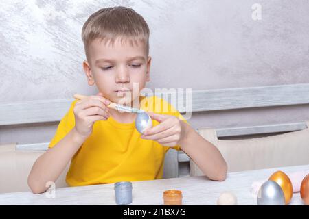 Ein kleiner blonder Junge malt Eier für die Osterferien in der heimischen Küche. Das Kind hat Spaß und feiert den Urlaub. DIY Ostereier Konzept. Stockfoto