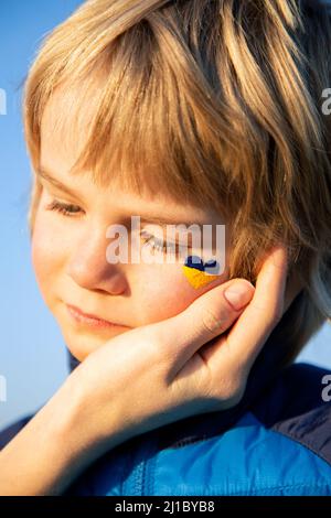 Nahaufnahme eines Jungen aus der Ukraine mit einem gelb-blauen Herzen auf der Wange und einer Frauenhand tröstet ihn sanft. Traurigkeit der kleinen Ukrainer Stockfoto