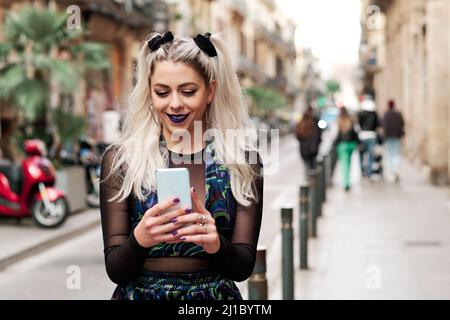 Alternative junge Frau, die ihr Mobiltelefon benutzt, während sie die Straße entlang geht. Stockfoto