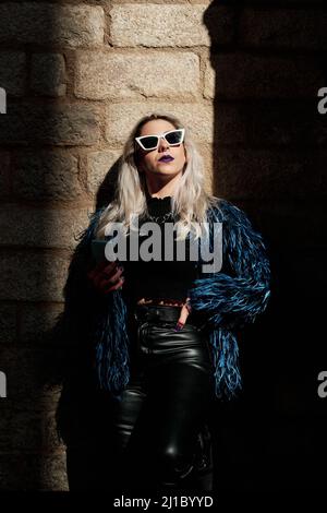 Eine alternative Frau in einer Sonnenbrille blickt auf die Sonne, während sie ein Mobiltelefon im Freien hält. Stockfoto