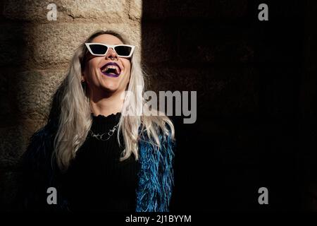 Alternative Frau in Sonnenbrille lacht, während sie draußen vom Sonnenstrahl beleuchtet wird. Stockfoto