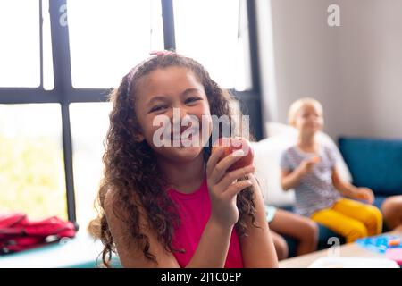 Porträt eines lächelnden biracial Grundschülerin hält Apfel, während auf der Couch während der Pause sitzen Stockfoto