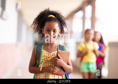Porträt eines lächelnden, biracial Grundschülerin, die Bücher hält, während sie auf dem Flur steht Stockfoto